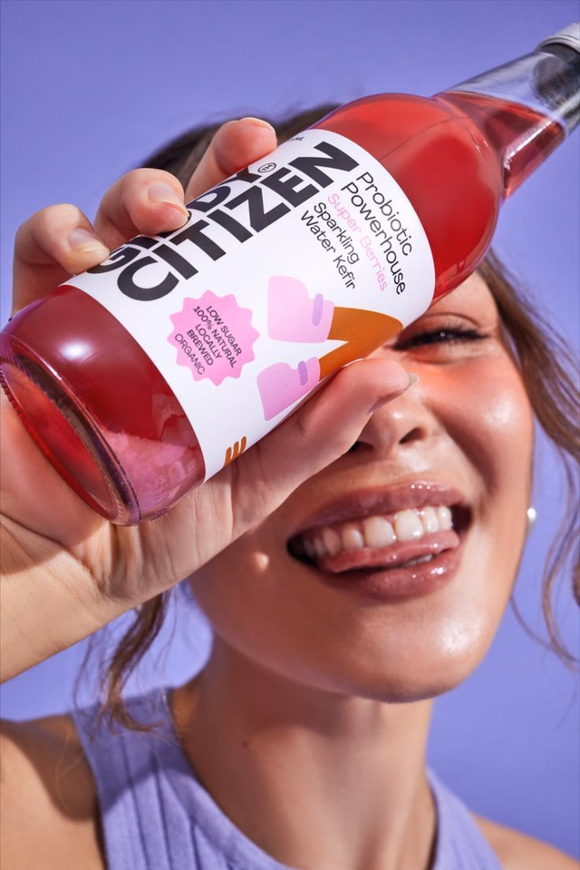 Smiling model holding a Citizen probiotic sparkling water kefir bottle on a purple backdrop